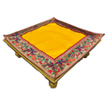 A square yellow woven mat or rug with an ornate embroidered border, displayed on a simple wooden stand against a plain white background.