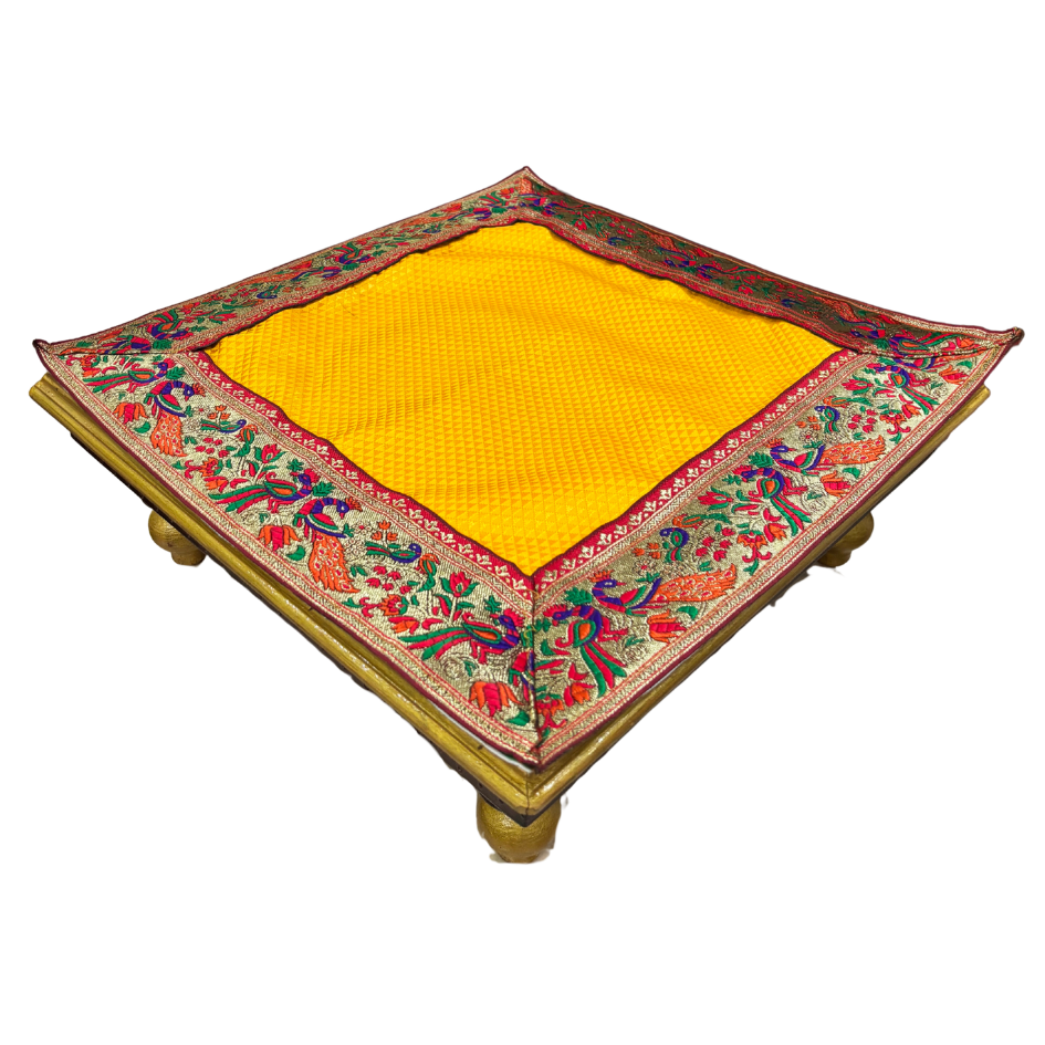 A square yellow woven mat or rug with an ornate embroidered border, displayed on a simple wooden stand against a plain white background.